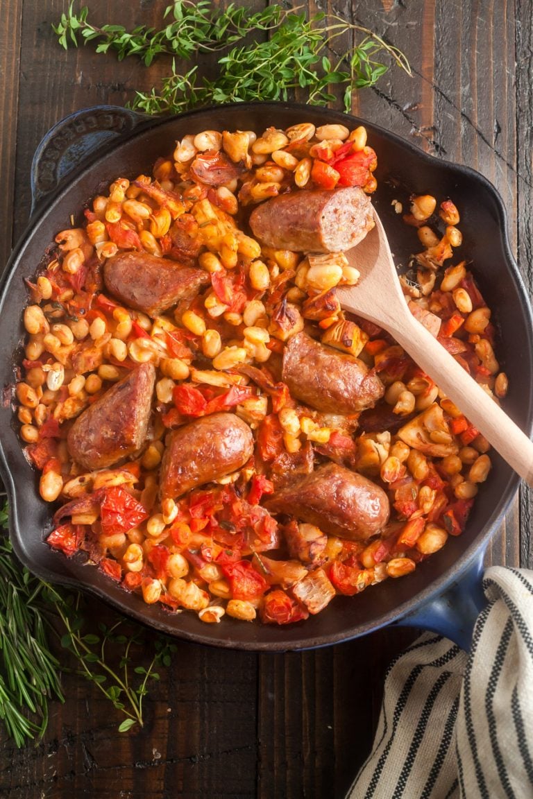an overhead view of turkey cassoulet in a pan, a wooden spoon removing a scoop of this white bean stew has a sausage link on it