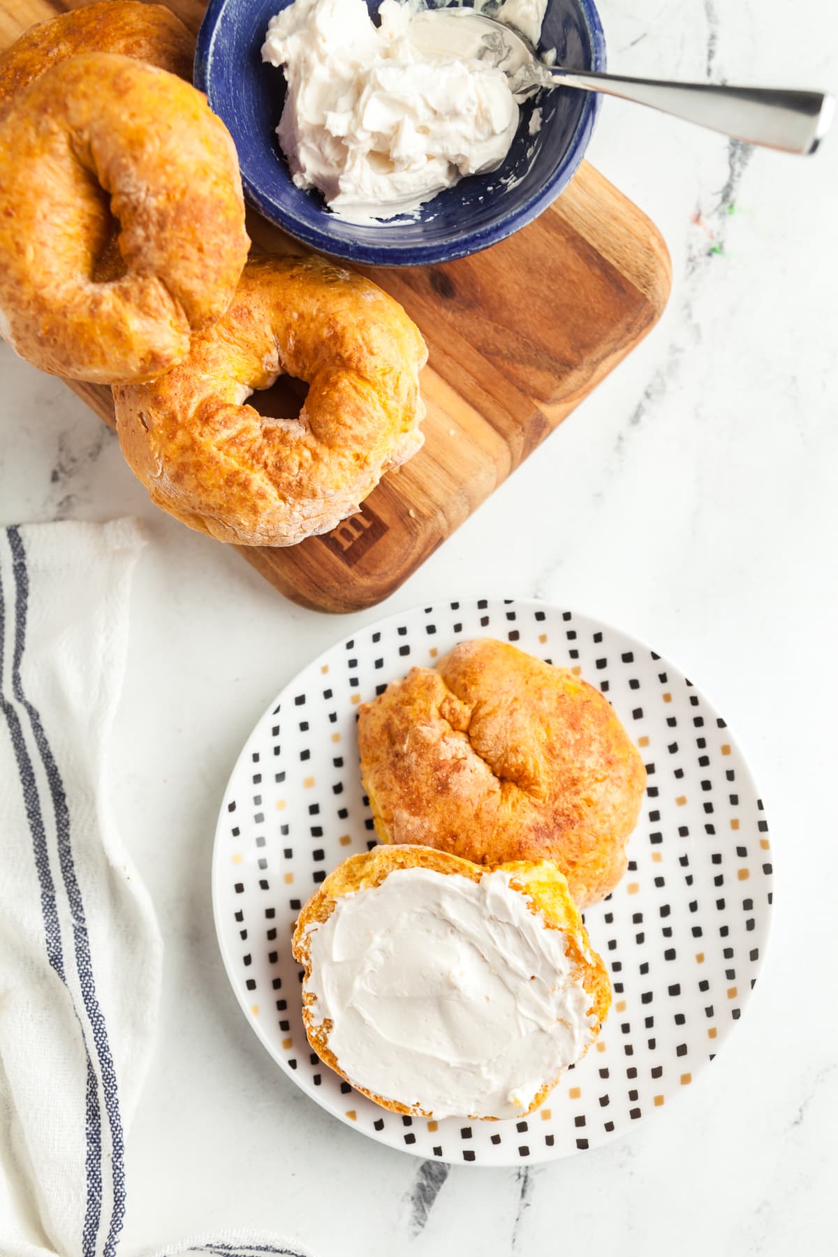 A small plate with a pumpkin bagel cut in half and slathered with cream cheese and a board of more air fried bagels in the back. 