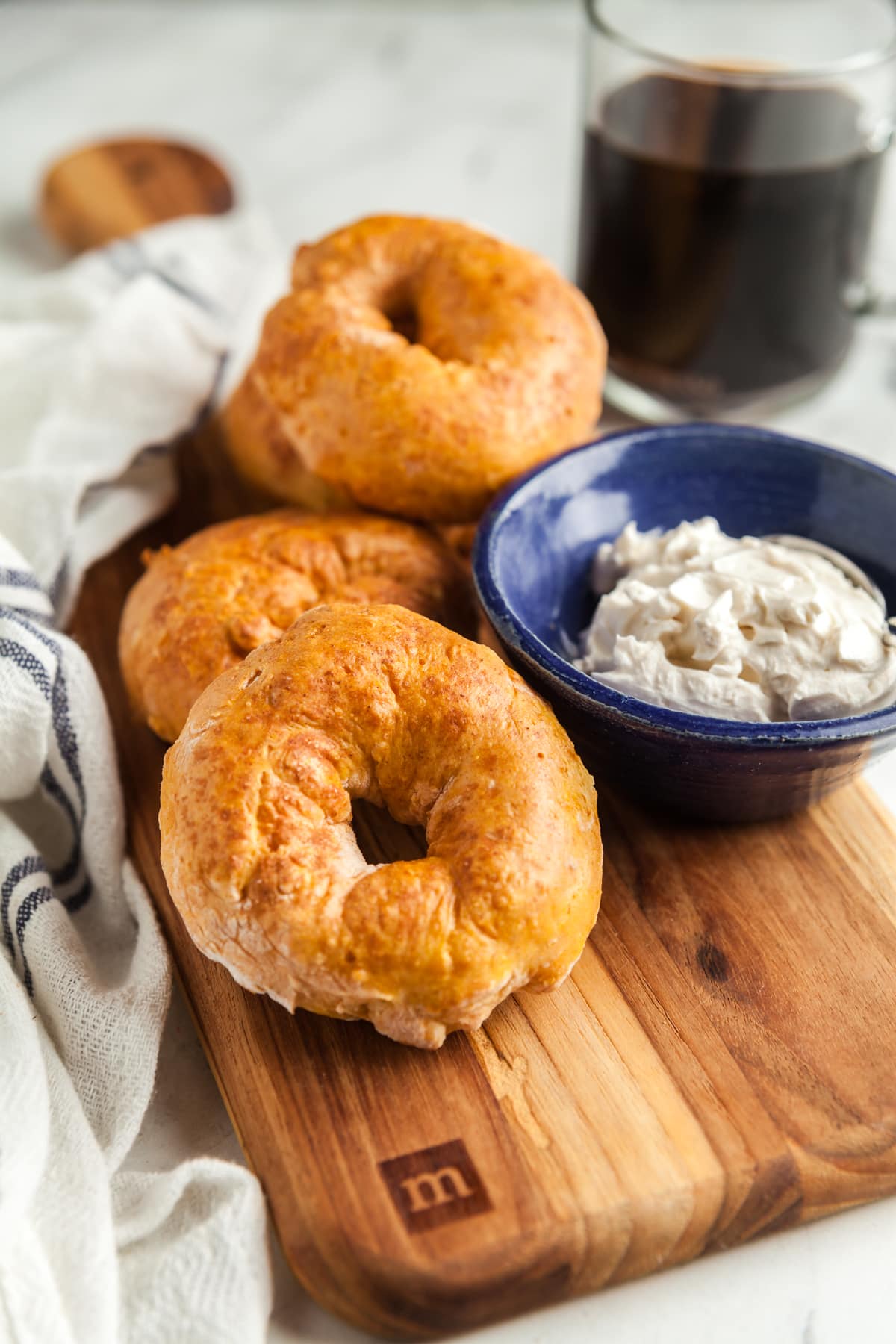 A wooden cutting board piled with baegls and a small blue dish of vanilla cream cheese with coffee in the background. 