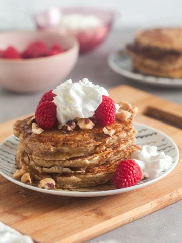 A stack of oatmeal pancakes topped with raspberries, hazelnuts, and whipped cream.