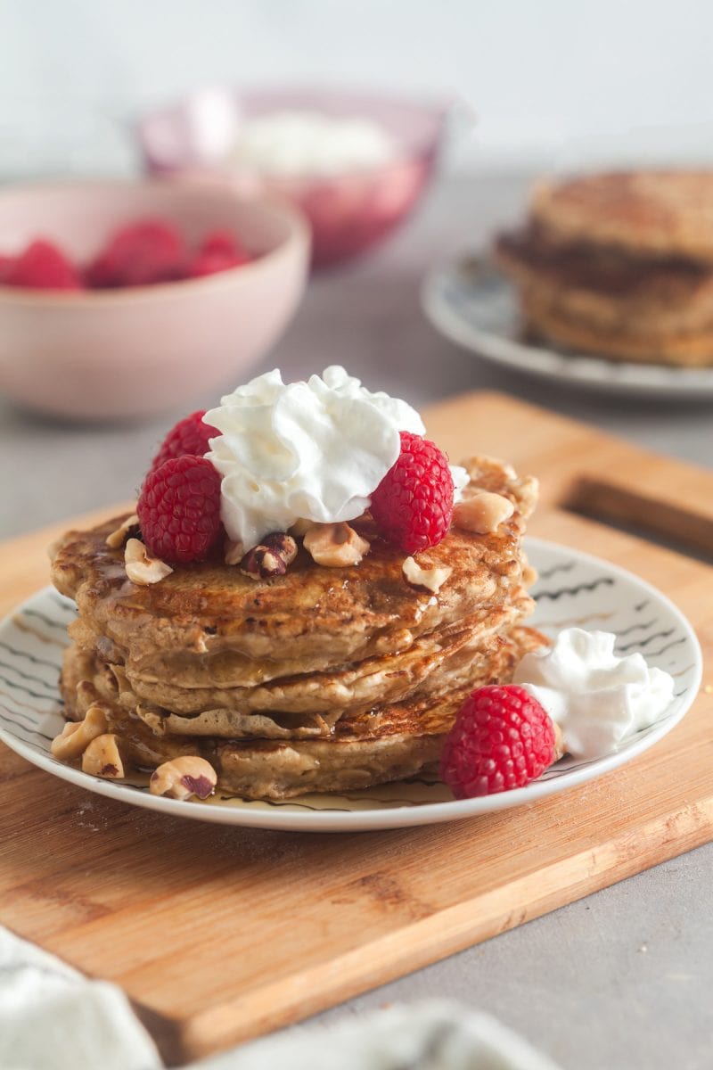 A stack of oatmeal pancakes topped with raspberries, hazelnuts, and whipped cream.