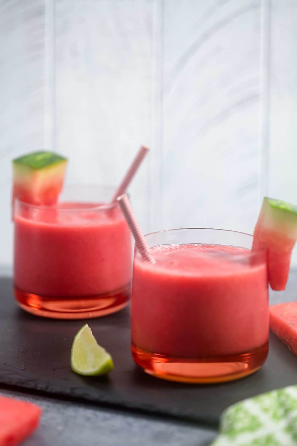Two 4-ingredient watermelon smoothies in short glasses on a dark grey slab and garnished with fresh watermelon. 