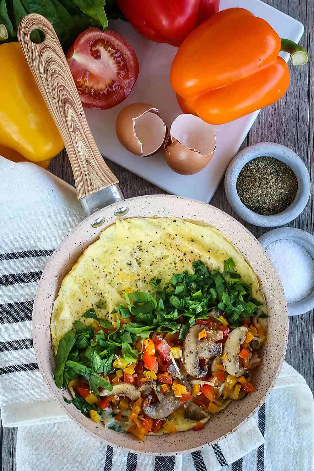 Overhead view of omelets with spinach in a pan surrounded by peppers and tomatoes