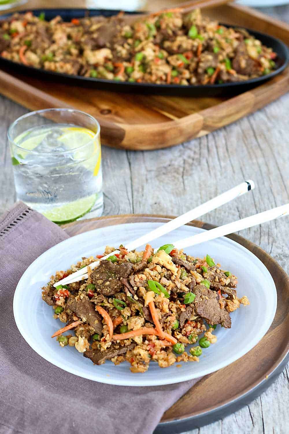 A white plate with beef stir fry and a drink on a table with a serving platter in the background.