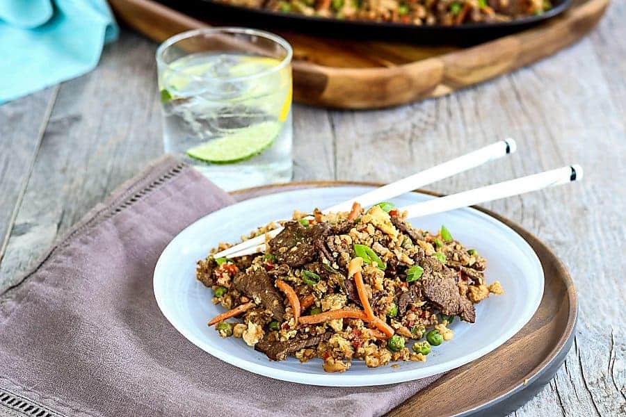 A white plate with beef stir fry and a drink on a table with a serving platter in the background.