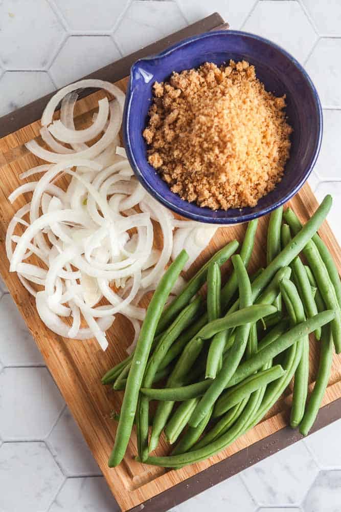 wood cutting board ion a white tile background with ingredients for low car and gluten-free green bean casserole: green beans, onions, and pork panko