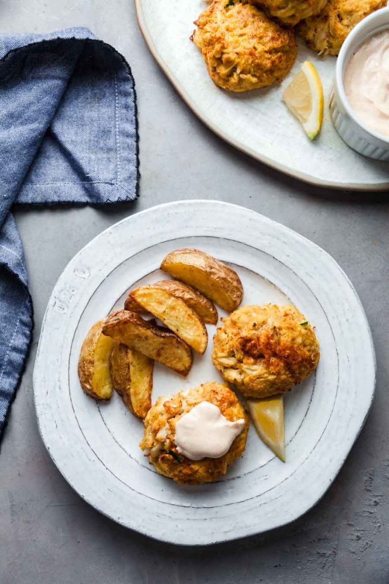 Overhead view of two crab cakes on a plate, with potato wedges on the side. One crab cake is topped with remoulade. 