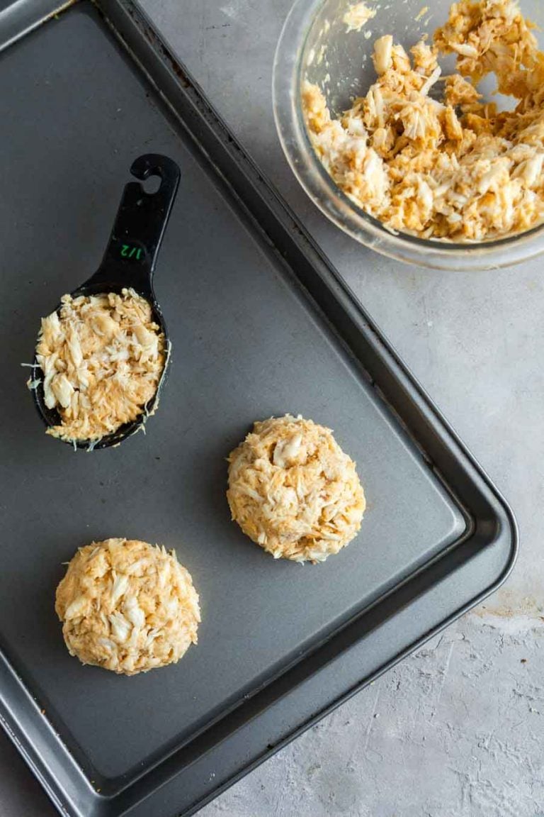 A sheet pan with uncooked crab cakes and a measuring cut filled with the crab mixture.