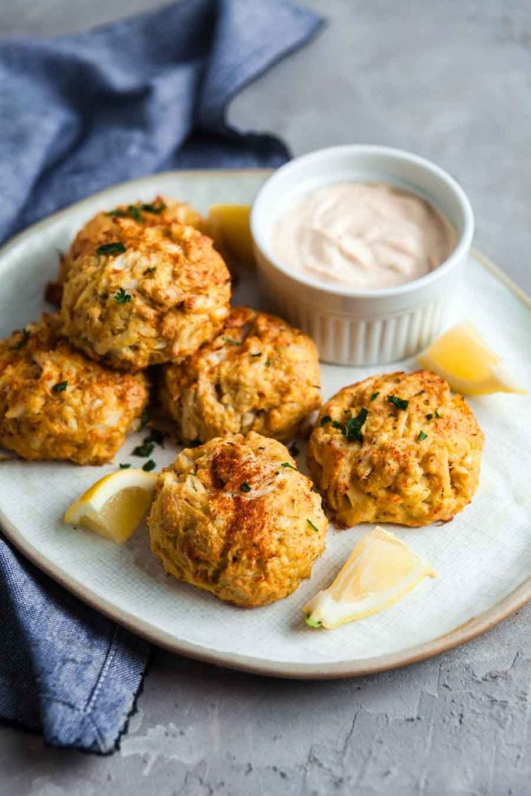 A white platter with 6 giant baked crab cakes and a small dish of remoulade on a grey background.