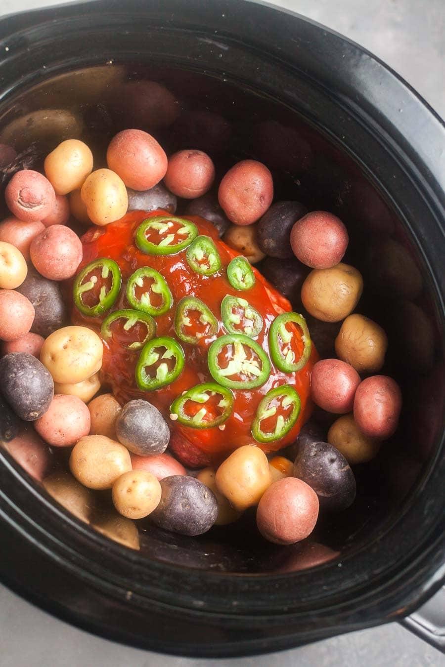 overhead view of the meatloaf and potatoes in a crock pot slow cooker. The meatloaf in the the center and is topped with homemade barbecue sauce and slices chile peppers. Small potatoes surround the meatloaf.