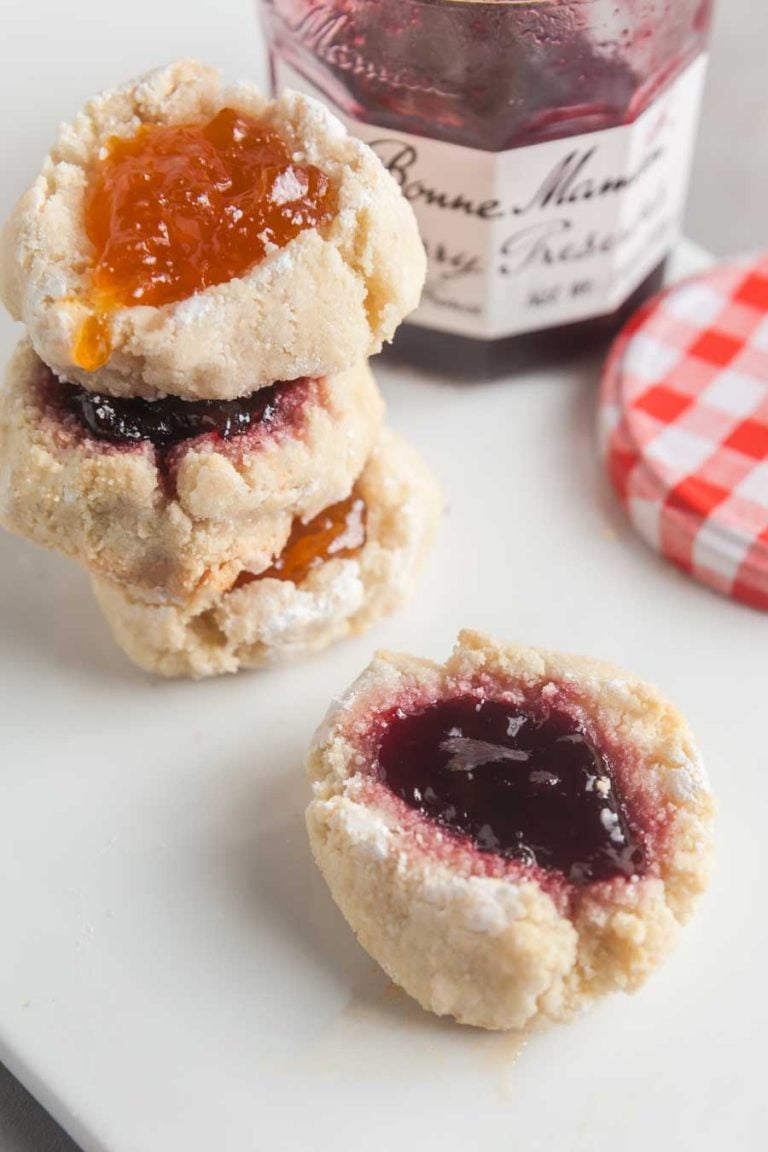 A stack of almond thumbprint cookies next to a jar or cherry preserves on a white background