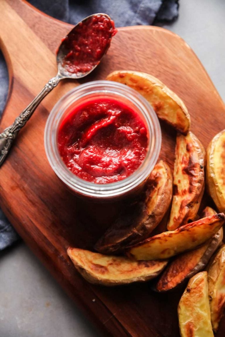 An overhead view of a jar of homemade ketchup with baked potato wedges and a spoon on a cutting board. 