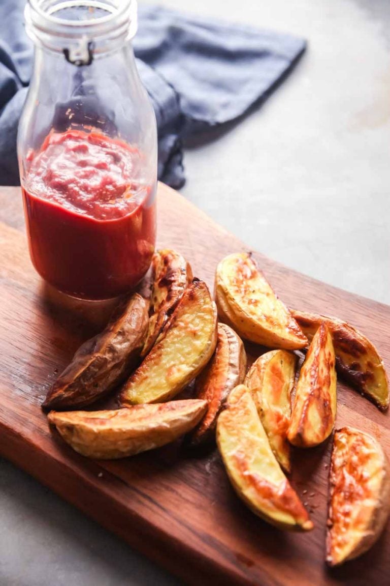 A tall jar of homemade ketchup on a wooden cutting board next to baked fries. 