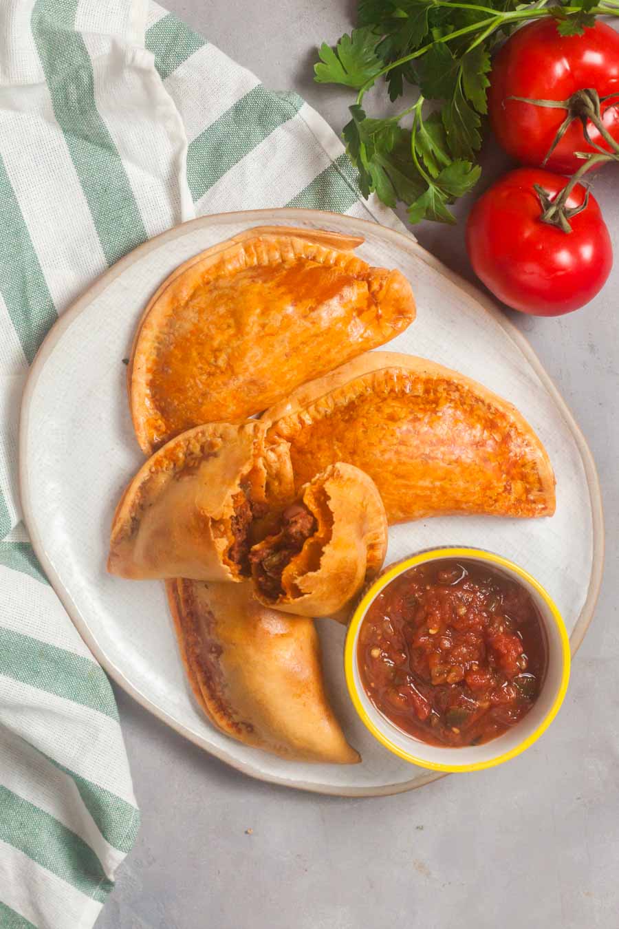 A white platter of air fryer empanadas with a dish of salsa. One empanada is broken open so you can see the chili filling inside. 