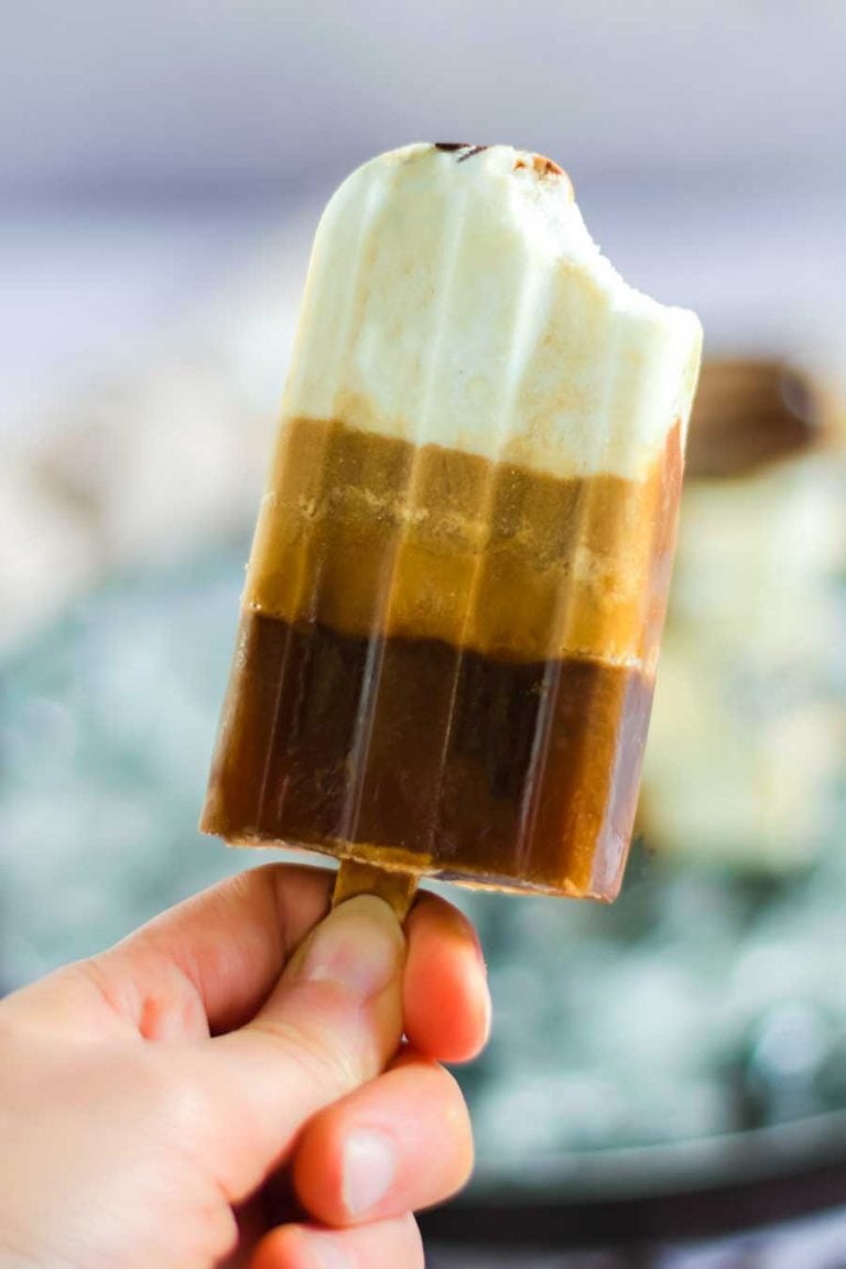 A closeup view of a coffee popsicle with a bite taken out. These ice pops feature three layers, a creamy chocolate chips ice cream layer, a smooth latte layer, and a bold coffee layer. 