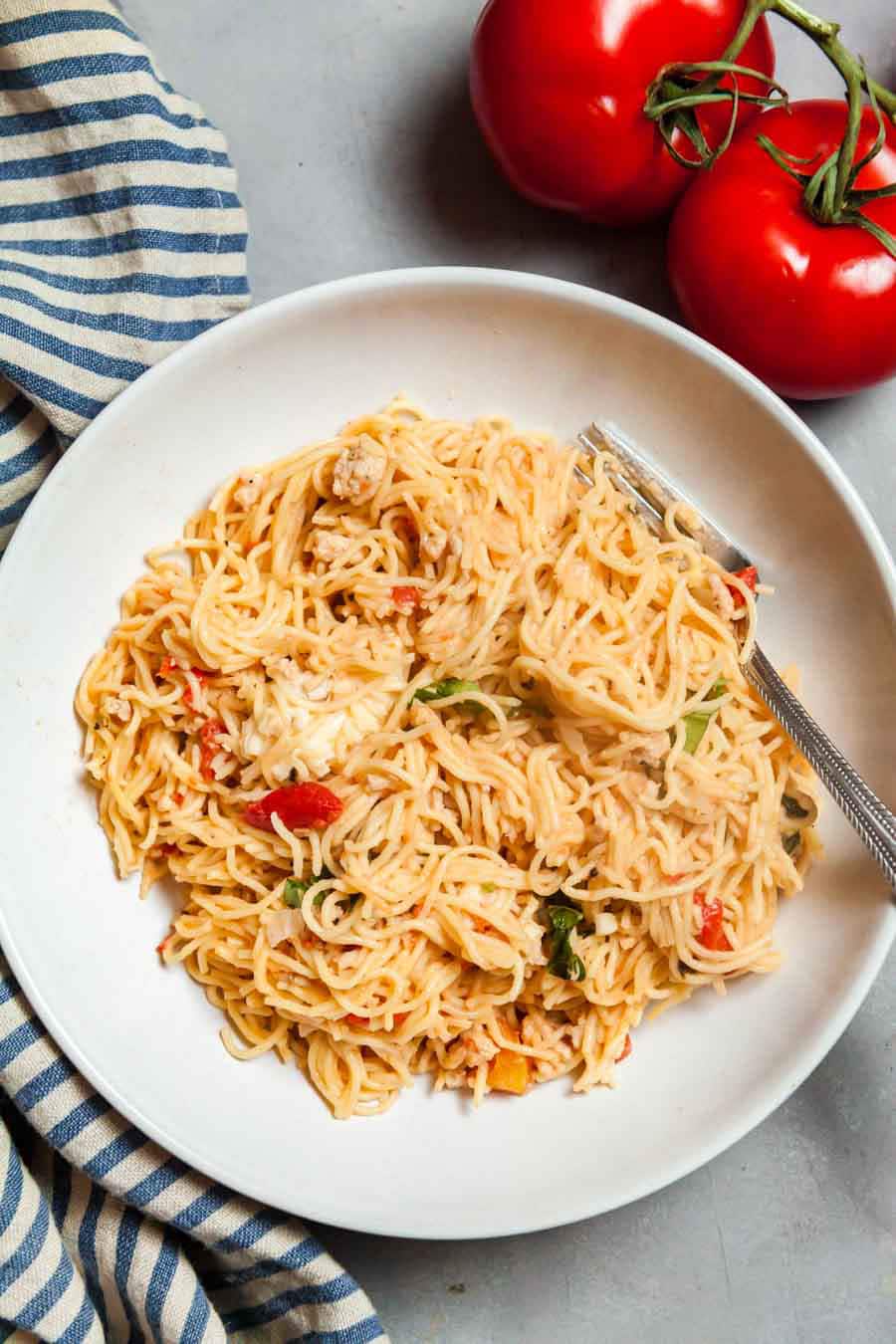 Overhead view of a white plate filled with chicken caprese pasta with a blue stripes napkin and red tomatoes. 