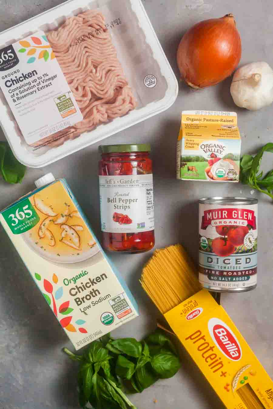 Overhead image depicting ingredients for making chicken caprese pasta include ground chicken, canned tomatoes, roasted red peppers, chicken broth, and fresh basil. 