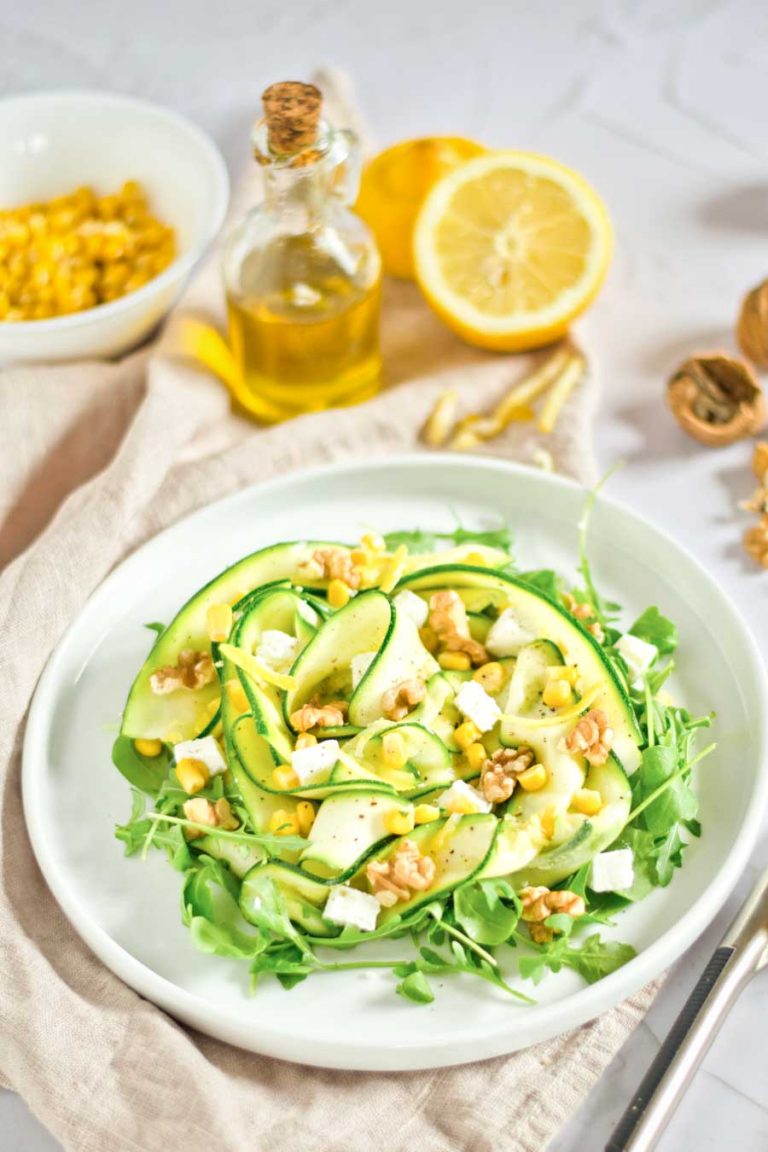 Zucchini and corn salad arranged on a white plate, with oil, lemons, and walnuts in the bacground. 