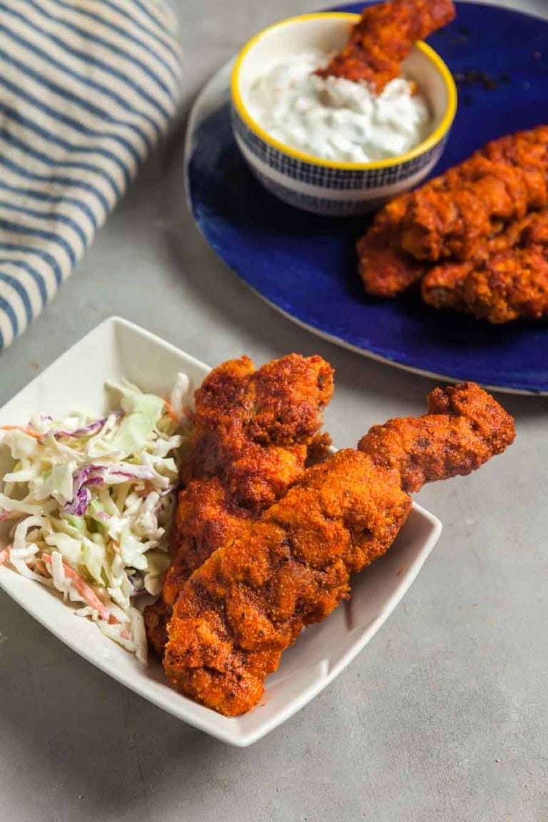 Two Nashville Hot Steak Fingers with coleslaw in a rectangular dish; a platter of steak fingers is in the background. 