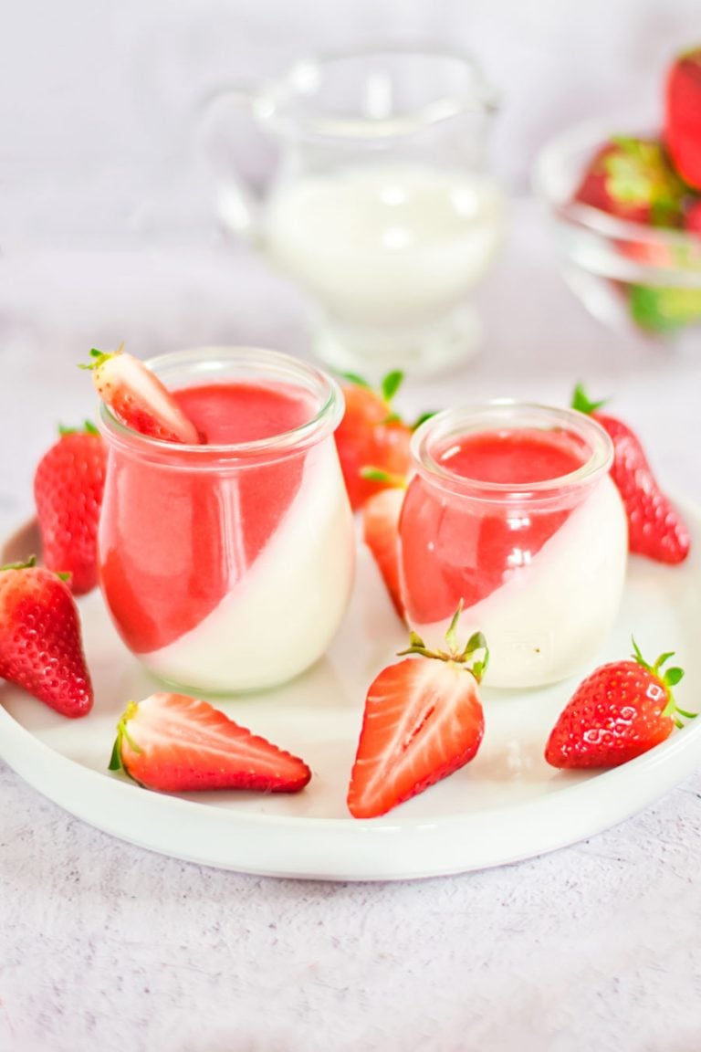 Two jars of strawberry panna cotta on a white tray, with ingredients and fresh strawberries in the background. 