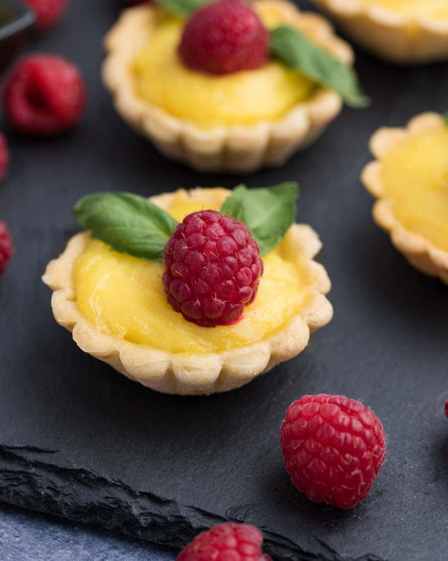 Miniature lemon tarts with fresh raspberries and mint on a slate board. 