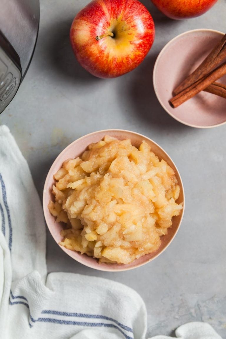 Overhead view of a bowl of applesauce on a grey tile backfrop with an instant pot, cinnamon sticks, apples and a tea towel in the background. 