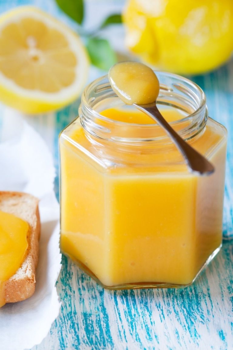 A glass jar of lemon curd on a blue background with cut lemons in the background. 