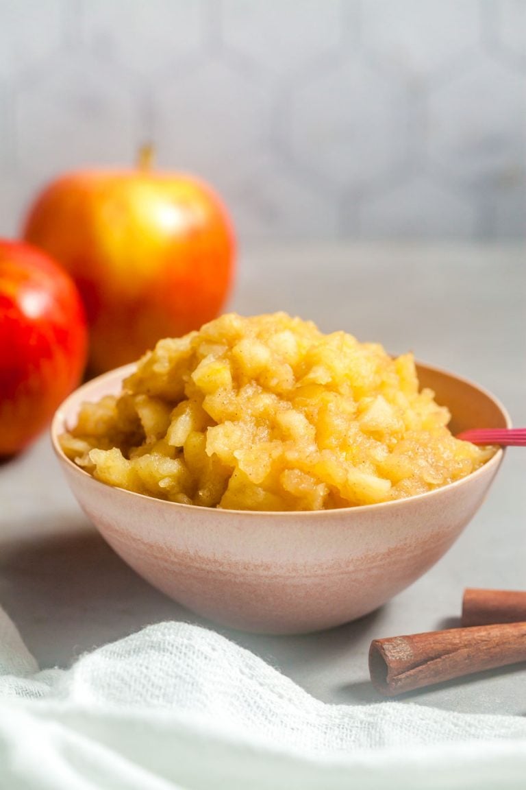 a small pink bowl of chunky applesauce in front of some apples, in front of a tiled wall. 