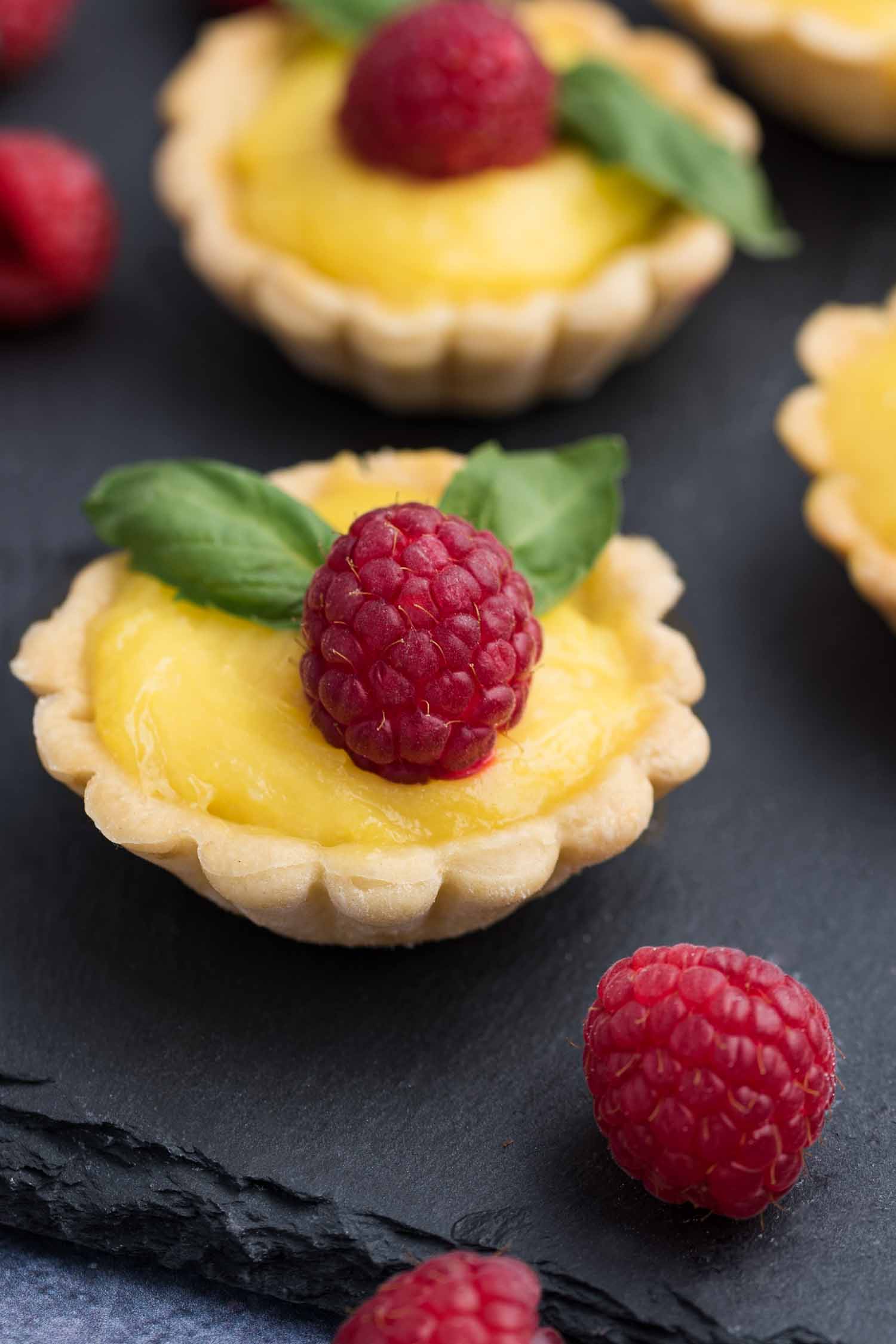 Mini lemon tarts topped with fresh raspberries on a slate board. 