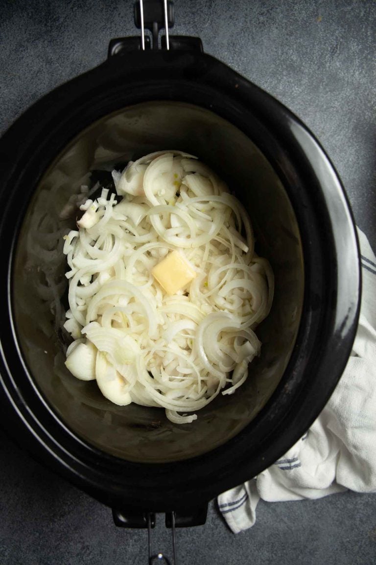 Overhead view of a slow cooker filled with raw sliced onions and apat of butter, ready to be transformed into slow cooker caramelized onions. 