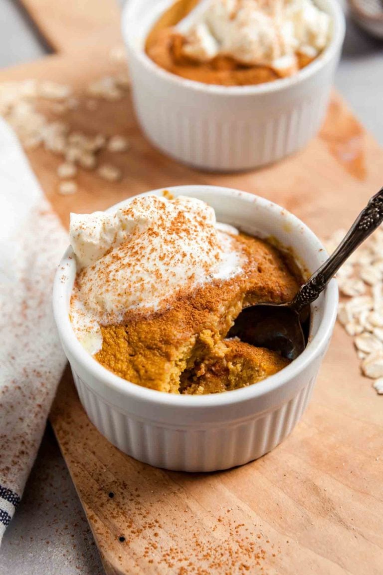 Two ramekins of pumpkin spice baked oats on a wooden board, with oats scattered around and a spoon digging in. 