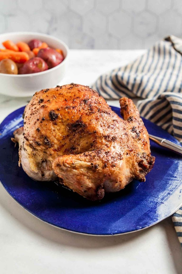 An air fryer rotisserie chicken with golden brown skin on a blue platter, with potatoes and carrots in the background. 