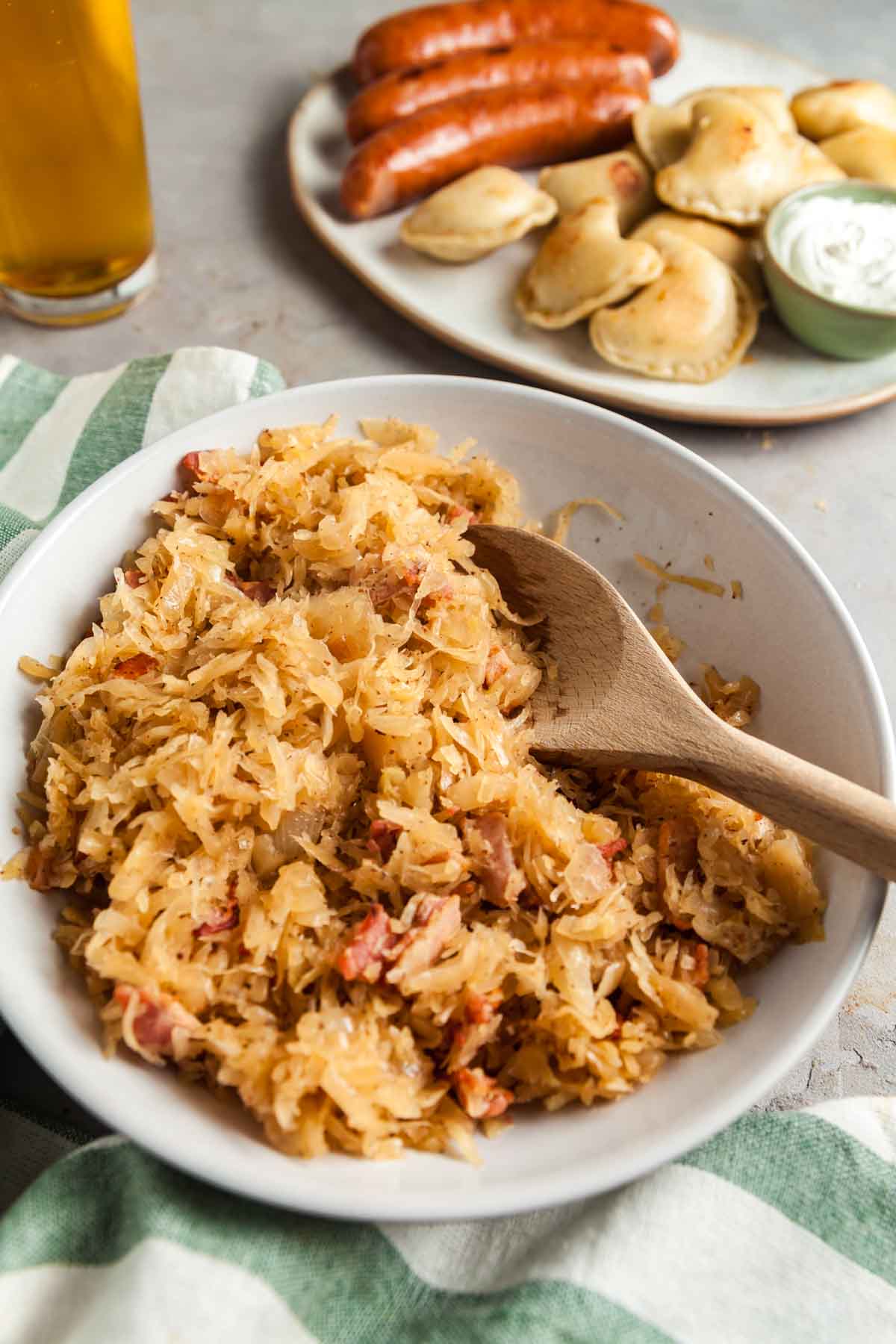 Closeup image of a serving bowl fileld with kapusta and a platter of pierogi and kielbasa in the background. 