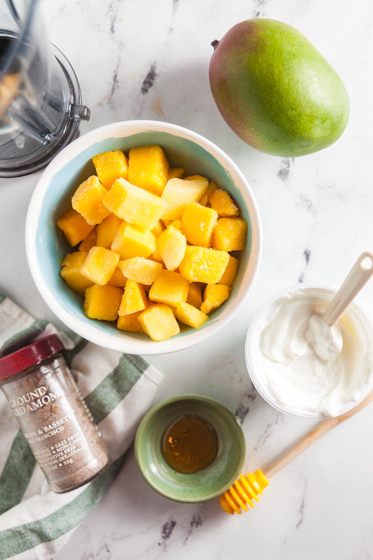 A flatlay image of ingredients needed ot make a mango lassi msoothie, arranged on a white countertop. Ingredients include frozen mango, yogurt, milk, and cardamom. 