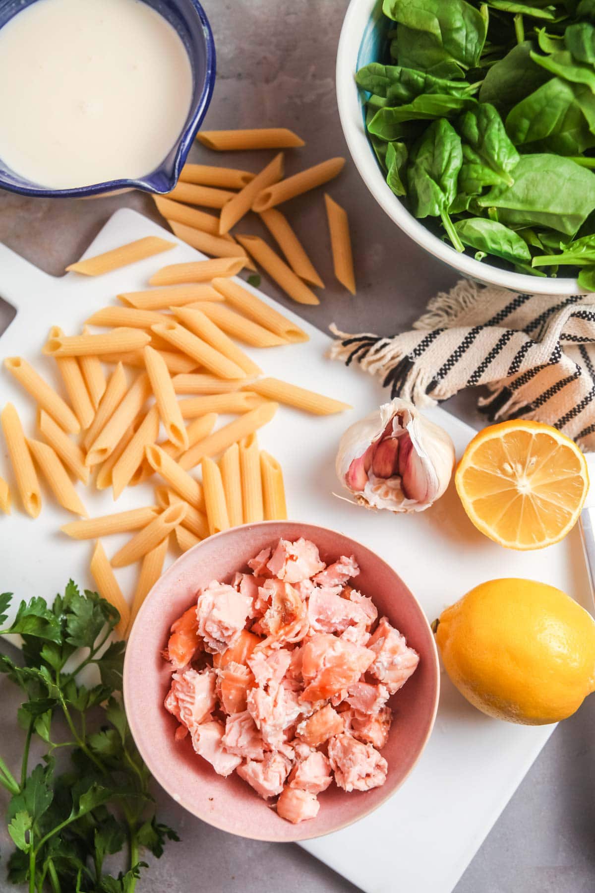 Overhead view of ingredients needed to make creamy salmon pasta: cooked salmon, penne, lemon, parsley, spinach, and heavy cream. 