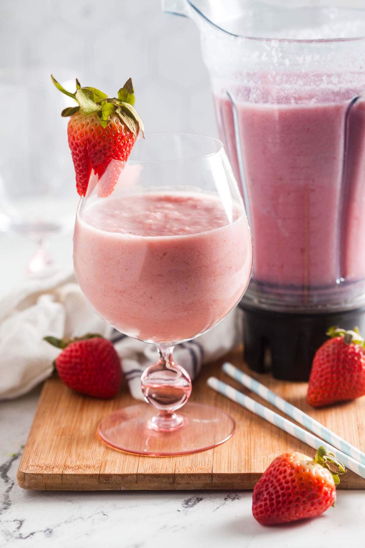 A goblet filled with strawberry banana smoothie and garnished with a fresh strawberry in front of a blender filled with smoothie. 