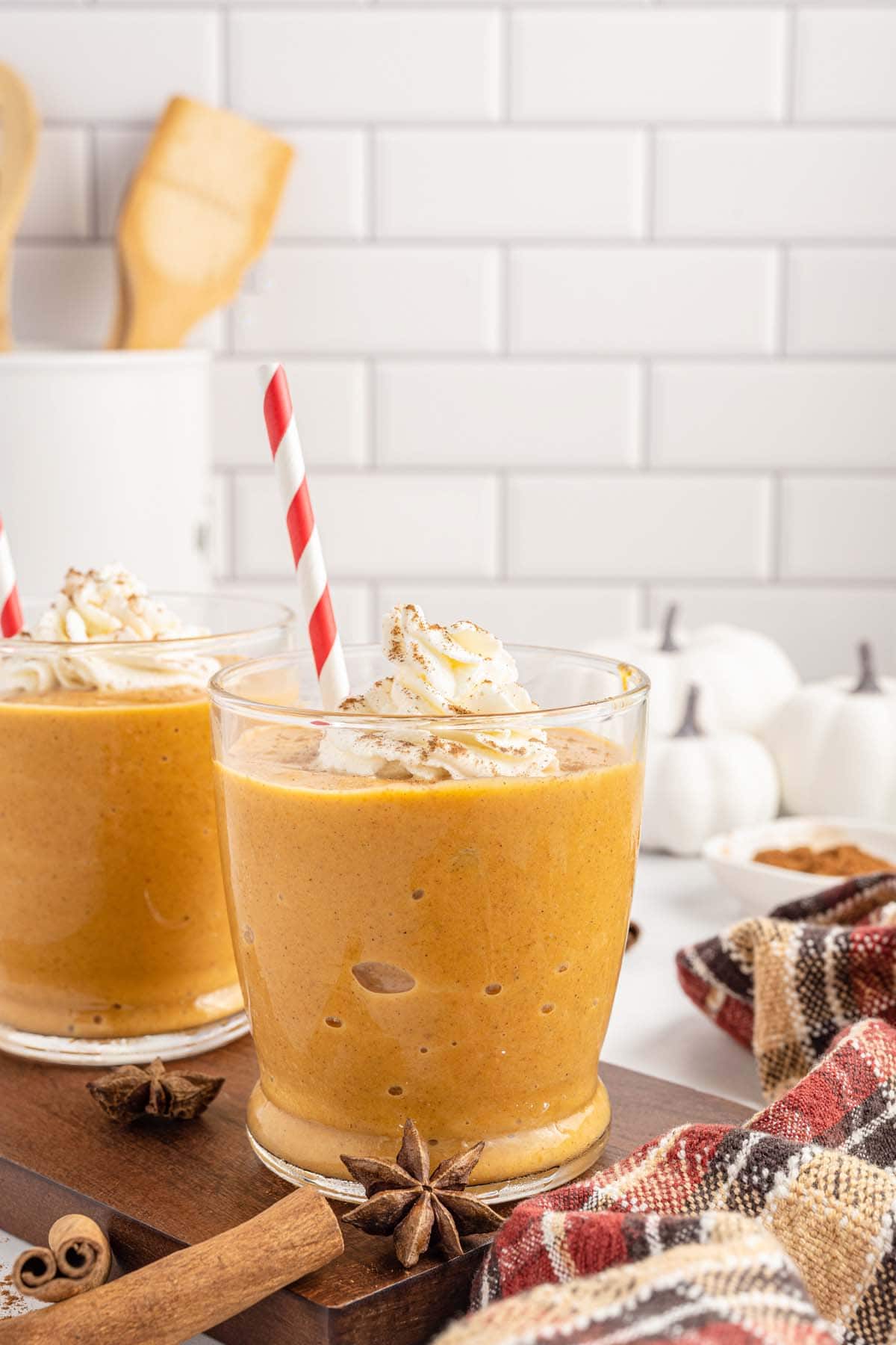 Two pumpkin pie smoothies topped with whipped cream in glasses with a red and white striped straw. 