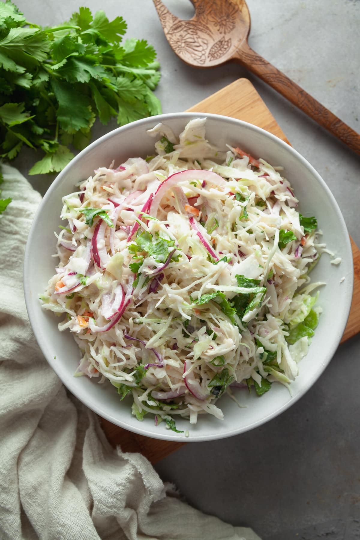 Overhead view of creamy coleslaw on a grey background, with a white linen and a wooden serving spoon. 