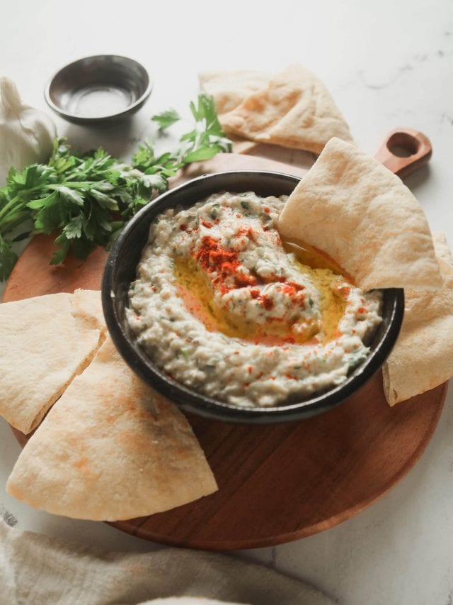 A cutting board with a bowl of baba ganoush and pita wedges.