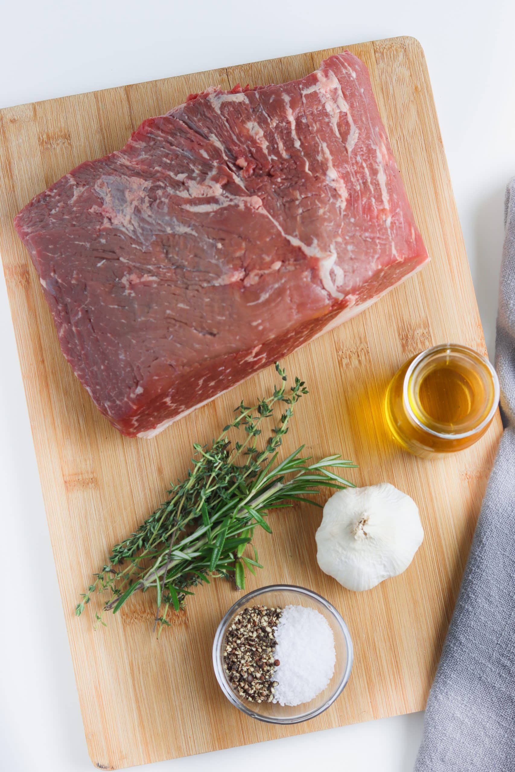 Flatlay image of ingredients needed to make a classic roast beef, including rump roast, fresh garlic and herbs, and olive oil. 