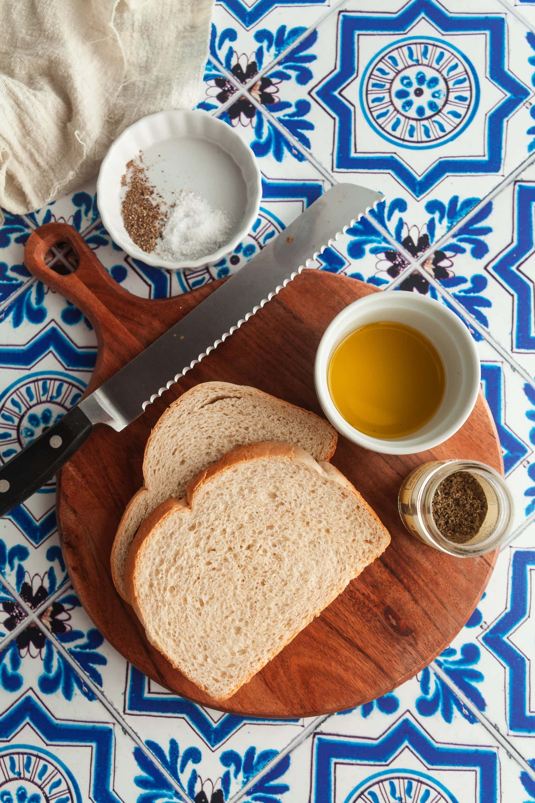 A wooden cutting board with sourdough bread, olive oil, seasoning, and salt on a blue tiled background. 