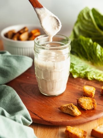 A glass jar of Greek yogurt Caesar salad dressing, with a spoon above it and homemade croutons and salad ingredients scattered around.
