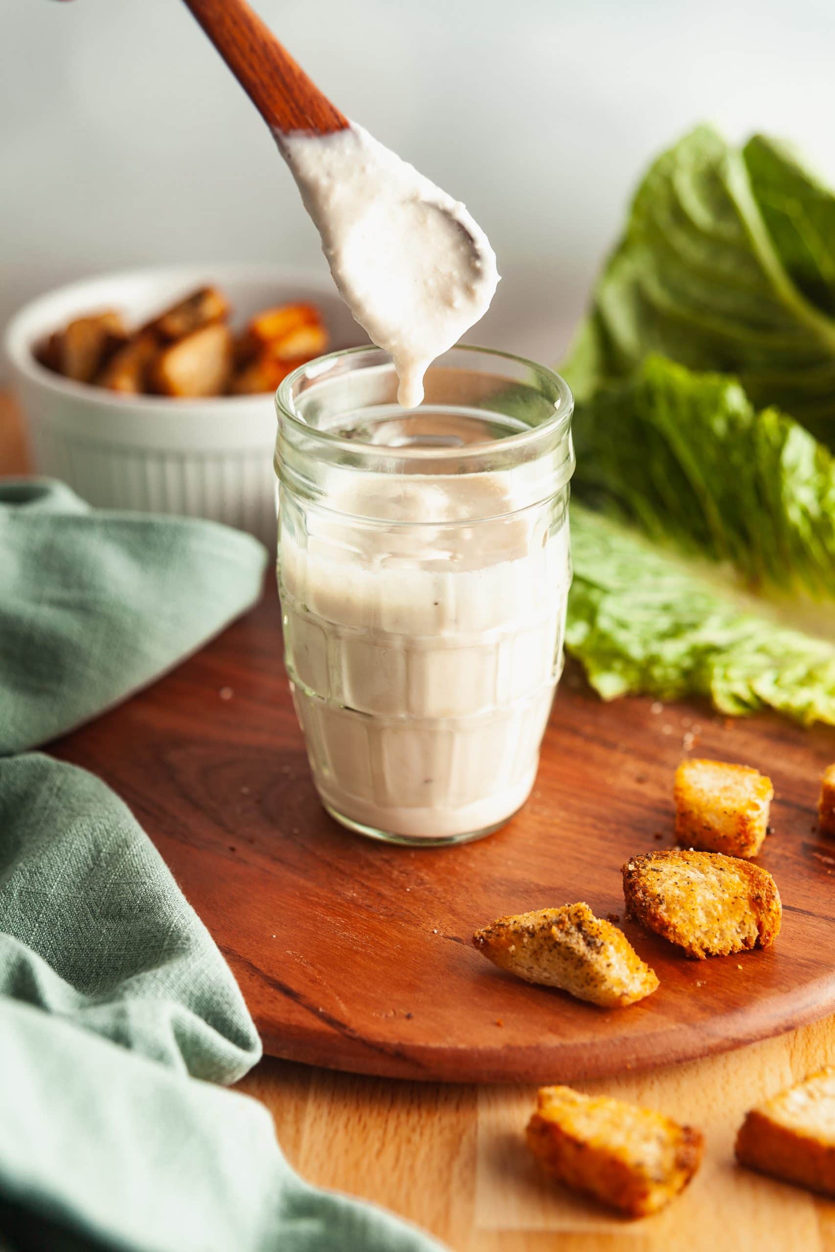 A glass jar of Greek yogurt Caesar salad dressing, with a spoon above it and homemade croutons and salad ingredients scattered around. 