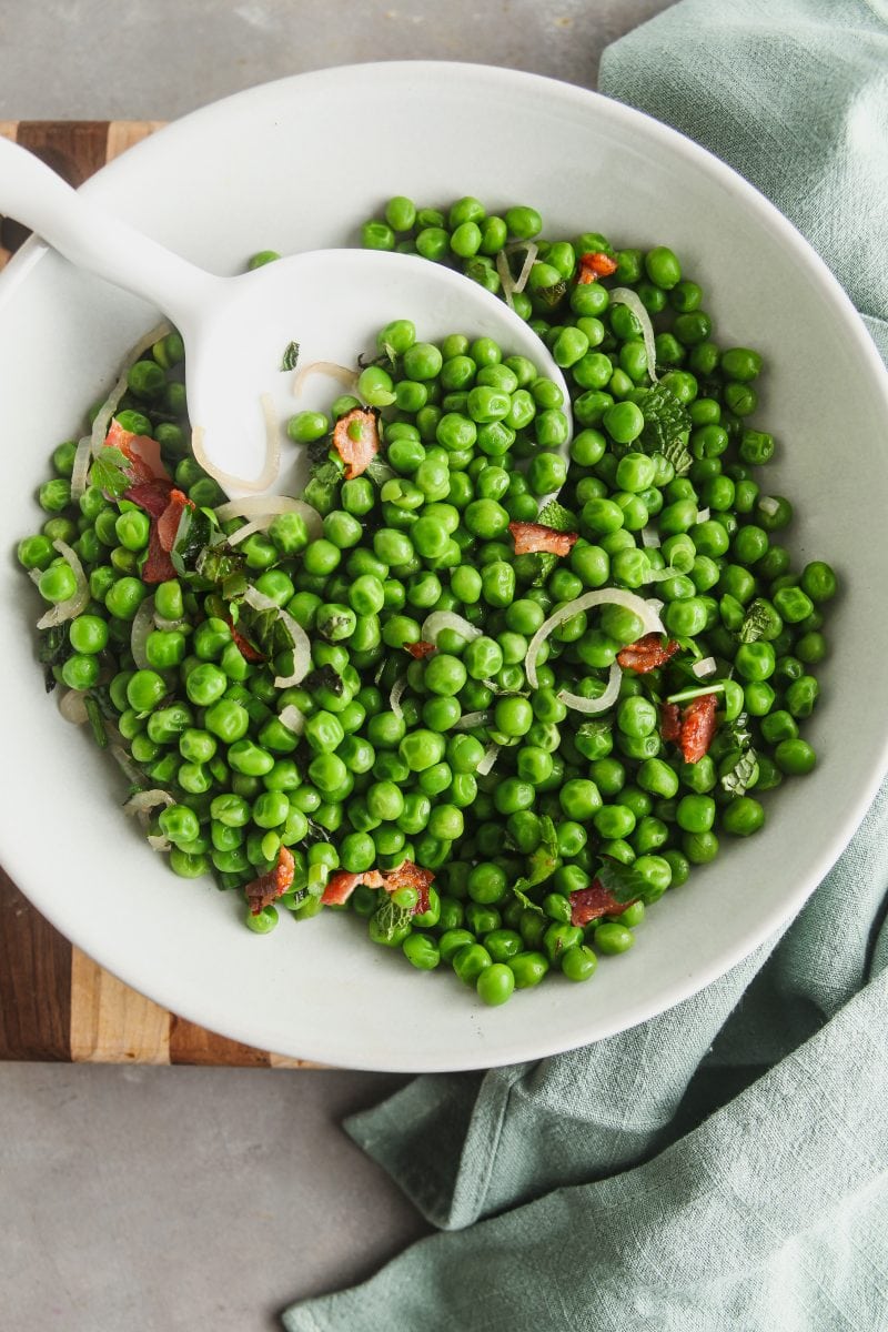 Close up image of peas with mint in a white bowl with a serving spoon.