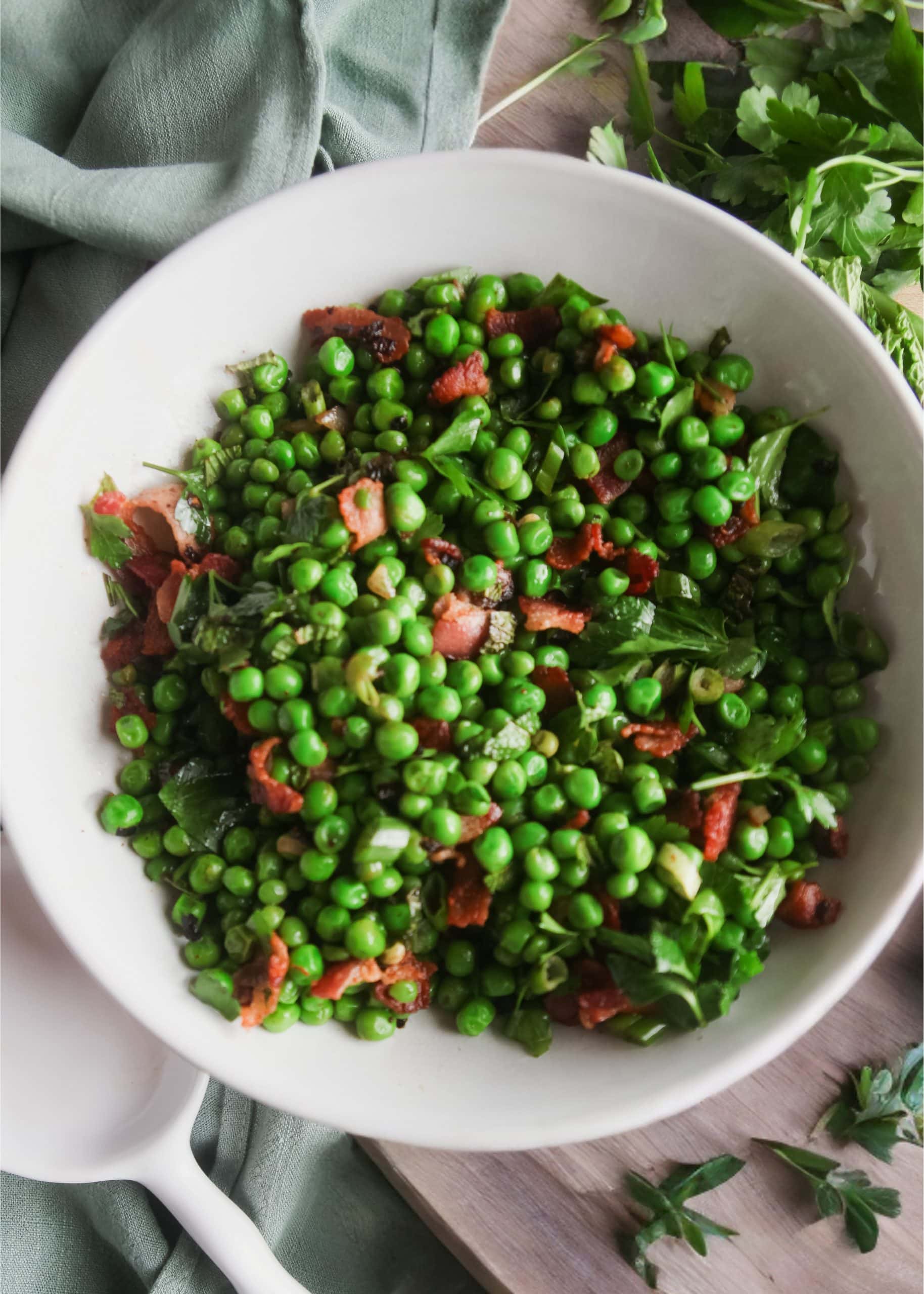 Overhead view of a white serving bowl filled with peas, bacon, and fresh herbs.